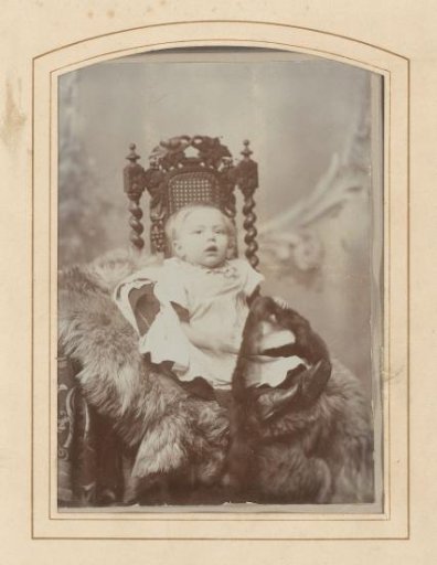 Portrait of a baby in a white dress with fur in hand, seated on a chair