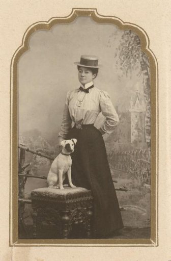 Portrait of a standing woman with a hat and a dog seated on a stool