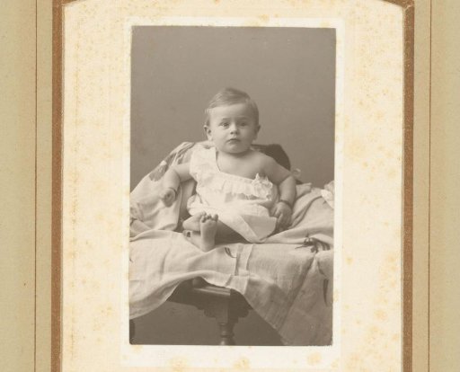 Portrait of a baby in a white dress, seated on a cloth