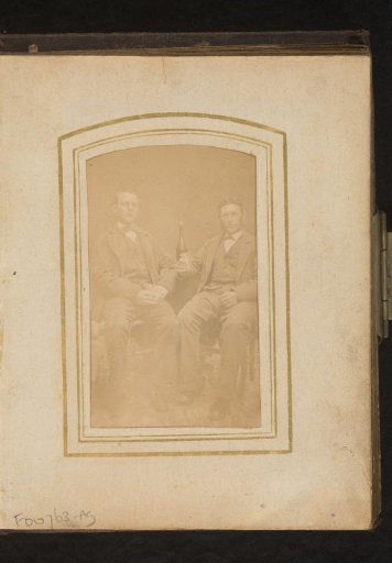 Portrait of two seated men with glasses in their hands and a bottle of wine behind them on a table