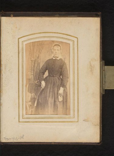 Portrait of a woman with a cap in the Overijssel folk costume, holding a book, standing by a chair