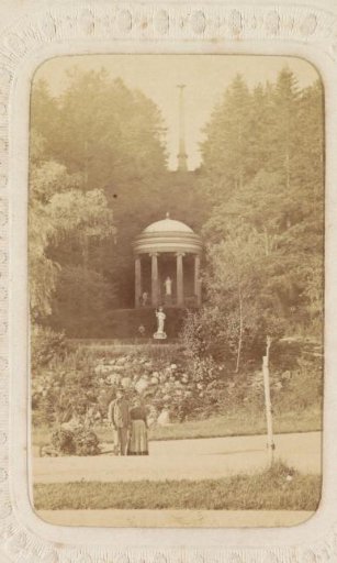 Man and woman posing in front of the tempietto in the Kleve gardens with in the background the obelisk commemorating the victims of the Franco-Prussian War in Kleve
