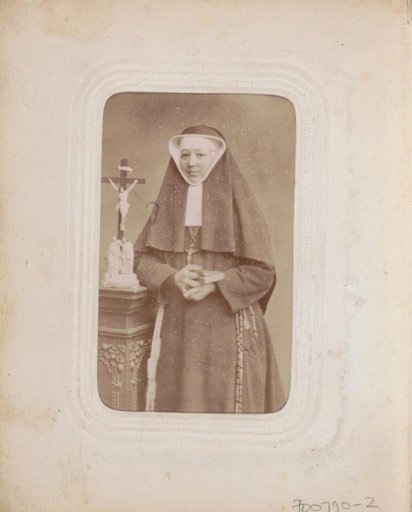 Portrait of a nun with a veil, rosary and crucifix around her neck, standing by a crucifix on a pedestal