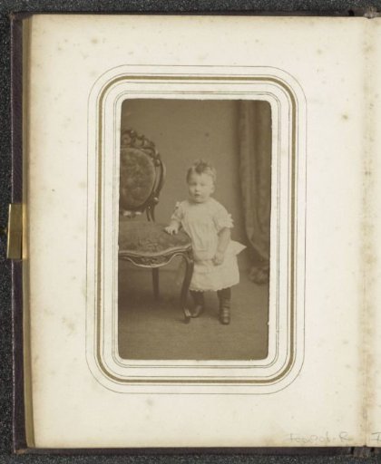 Portrait of a toddler in a white dress standing by a chair