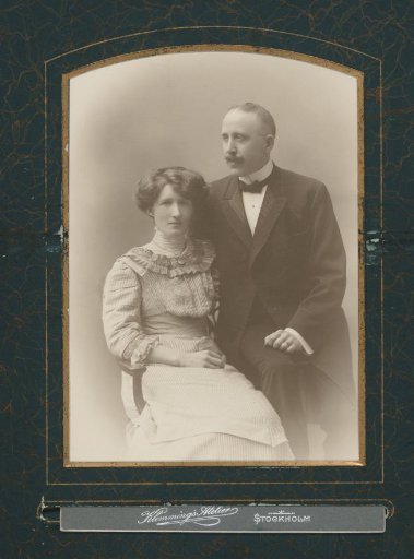 Portrait of a seated woman and a man, seated on the back of a chair