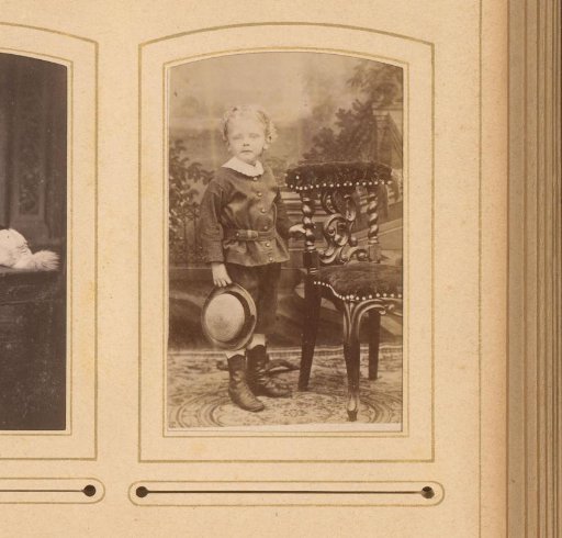 Portrait of a boy with a hat in his hand, standing by a chair