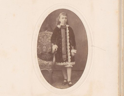Portrait of a girl in a jacket over lace trousers, standing by a chair