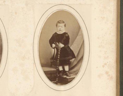 Portrait of a child with a hat in his hand, standing by a chair