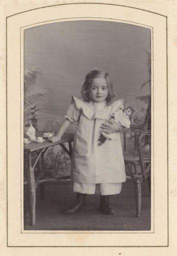 Portrait of a girl holding a doll, standing by a table with a tea service