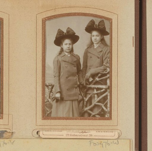 Portrait of two girls with cloaks and hats with bows, standing by a fence