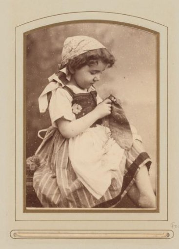 Portrait of a seated girl sewing a sock, in costume with apron and scarf on her head