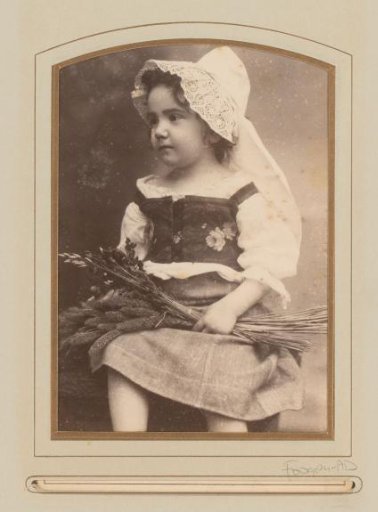 Portrait of a seated girl in traditional dress with a white cap, holding ears of corn