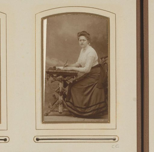Portrait of a woman with spectacles, seated at a table with a pen in her hand