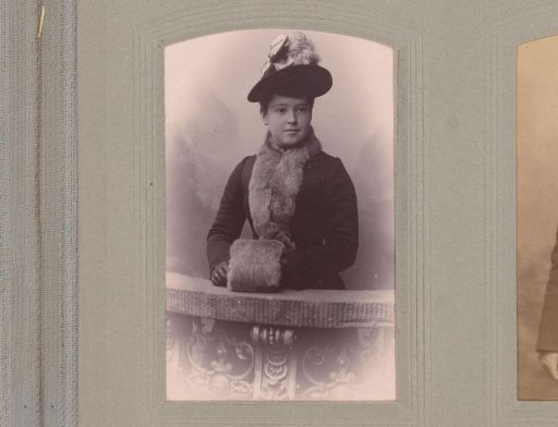 Portrait of a young woman with a coat, hat, and muff, standing by a balustrade