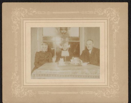 Portrait of a man and two women in traditional dress, seated at a kitchen table