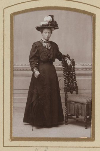 Portrait of a woman with a hat, standing by a chair