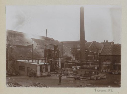 Factory grounds with buildings, a chimney, wooden barrels, and a worker