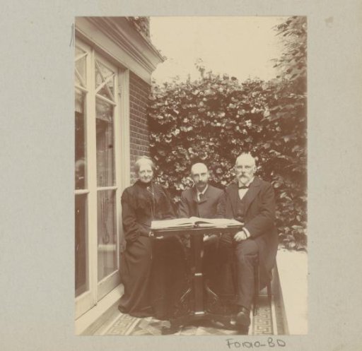 Two men and a woman sitting by a house with a book on the table
