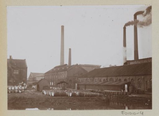 Exterior of factory buildings and smoking chimneys, with wooden casks in the foreground