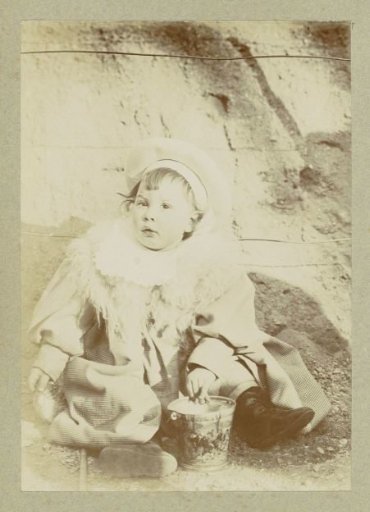 Portrait of a young child with a jacket, a scarf and a cap, sitting by a toy bucket