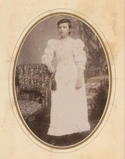 Portrait of a young woman in a white dress, standing by a chair