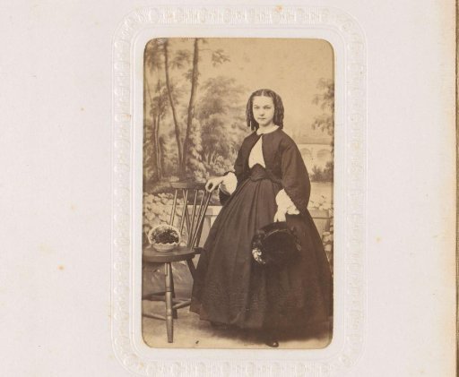 Portrait of a young woman with a hat in her hand, standing by a chair