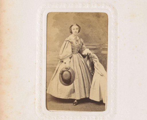 Portrait of a young woman with a hat in her hand, standing by a chair on which a cloak lies