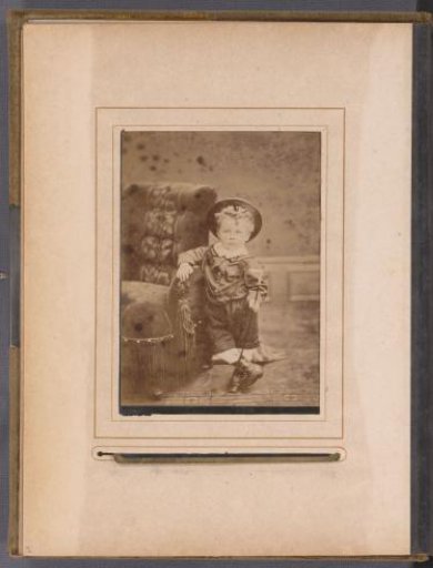 Portrait of a boy with hat and walking stick, standing by a chair