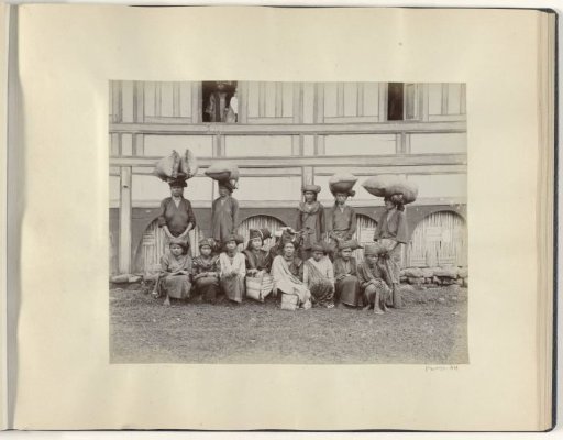 Women sitting and standing in front of a house with bales on their heads in the Dutch East Indies
