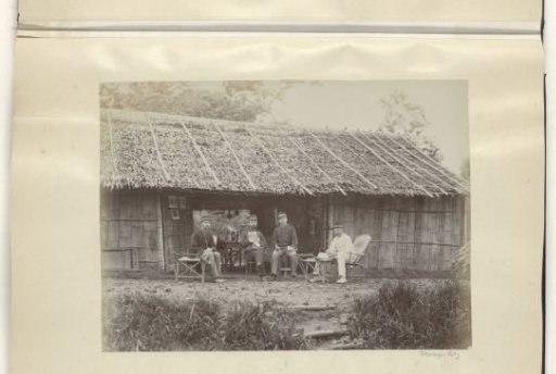 Dutch soldiers sitting in front of a wooden house in the Dutch East Indies, one reading a Breda ('Bredasche') newspaper