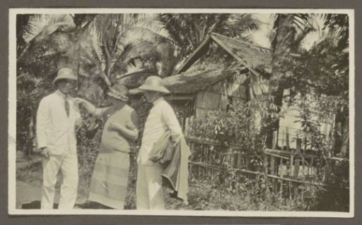 Two men in tropical clothing and a woman by a hut