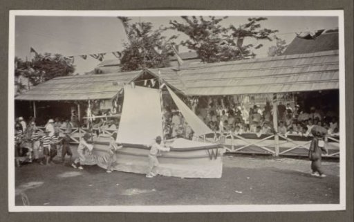 Participants in a parade pushing a pageant wagon with a sailboat