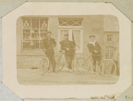 Three men with bicycles in military costume in front of a shop, probably in France