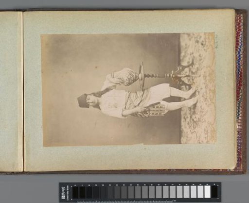 Portrait of a girl with fez and three-quarter trousers, standing by a guéridon with a vase
