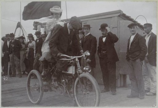 Race driver De Perigord, participant in the Paris-Amsterdam race, on his motorcycle with a woman behind him, presumably at Nijmegen