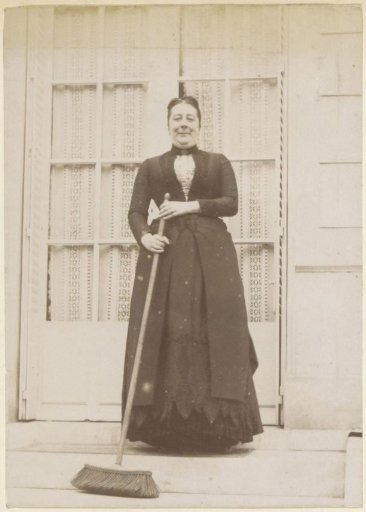 Portrait of a French woman, standing with a broom in her hand on the pavement in front of a house