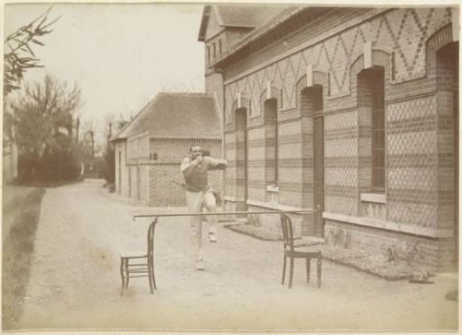 French man jumping over a stick between two chairs in a yard