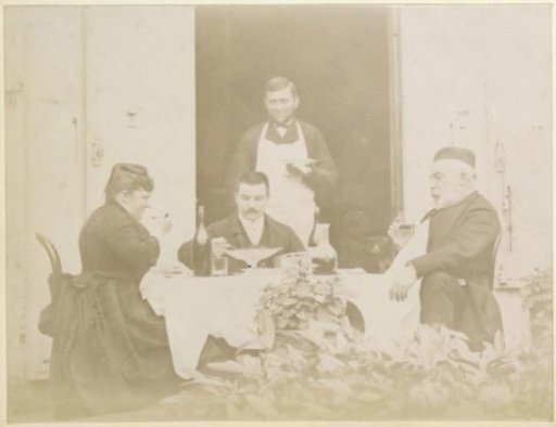 Two men and a woman dining at a table in France, the cook in the doorway