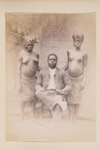 Studio portrait of a Western-dressed African man with two half-naked women, probably in Cameroon