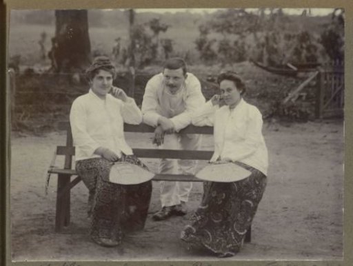 Two seated women and a man leaning on a bench, possibly in Suriname