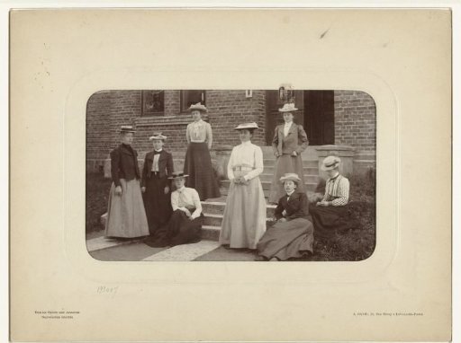 Group portrait of eight unknown women on a porch, all wearing hats