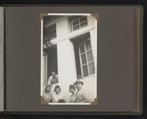 Three Women and a Man Standing in Front of a House, Behind Them a Smoking Man and a Dog Lean Out of the Windows