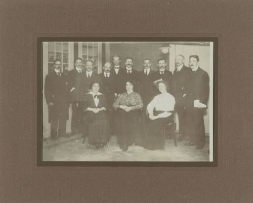 Group photograph of three seated women with a group of standing men in suits behind them