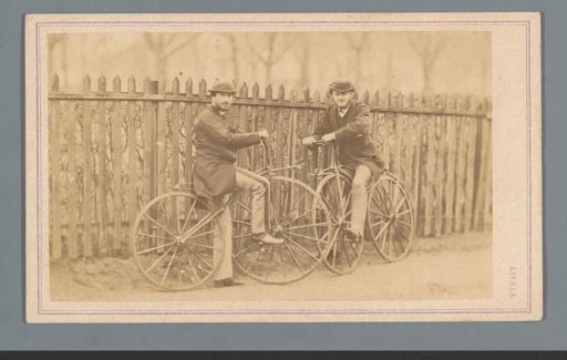 Two unknown men on bicycles in front of a fence