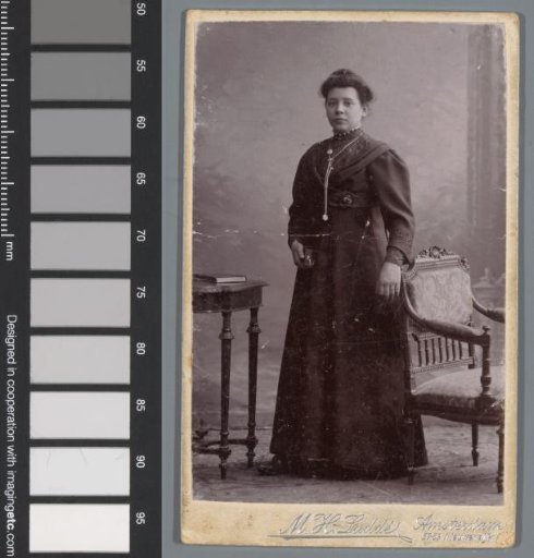 Portrait of an unknown woman, standing between a table and an armchair
